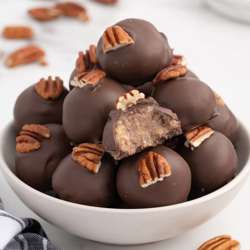 A bowl of condensed milk pecan balls coated in chocolate, topped with pecans, with one ball showing the creamy filling.