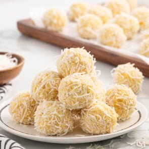 A plate of pineapple balls coated in shredded coconut, with more arranged on a wooden tray in the background.