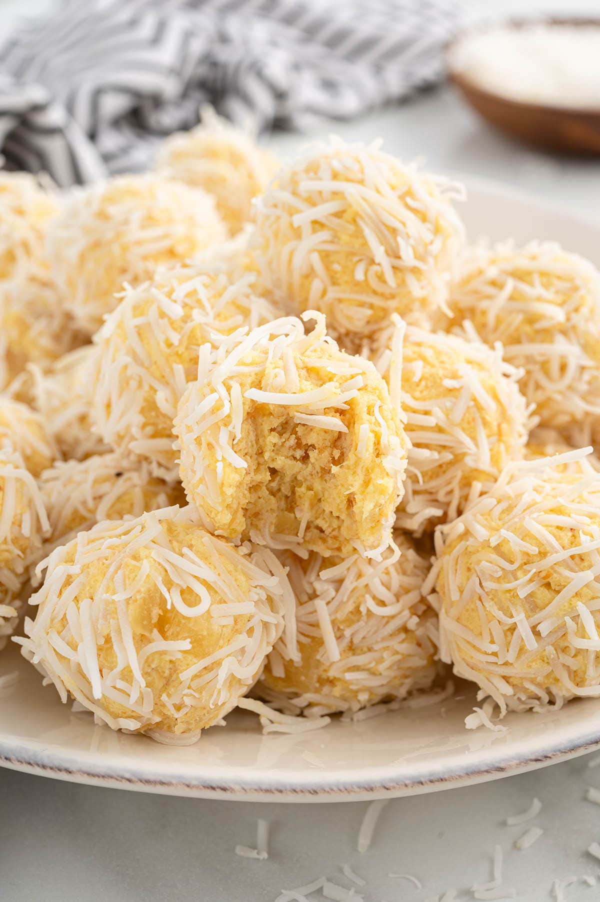 Pineapple balls coated in shredded coconut, piled on a plate, with one bitten piece. 