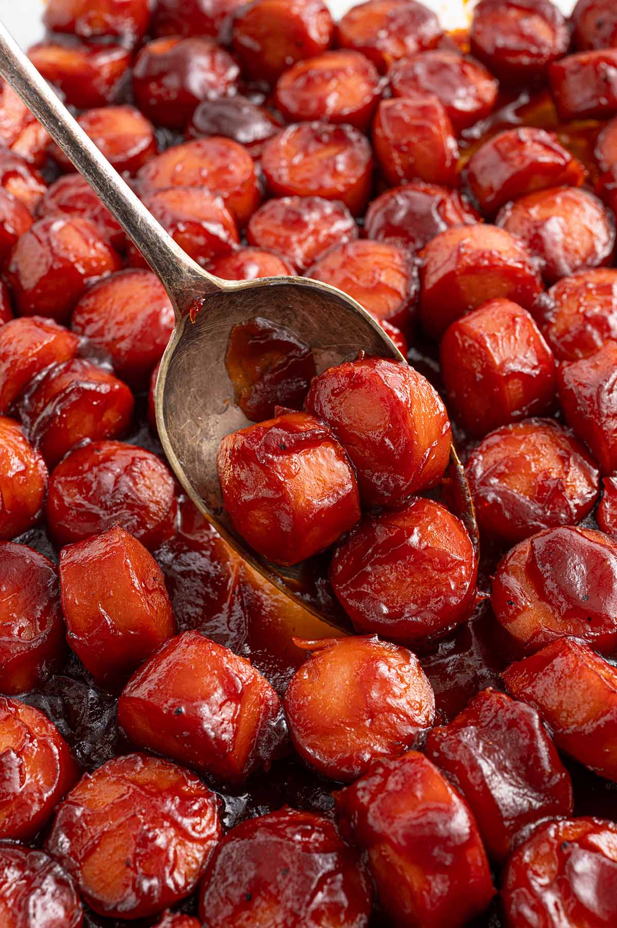 Close-up of juicy sausage bites coated in a thick, sticky barbecue glaze, being served with a spoon.