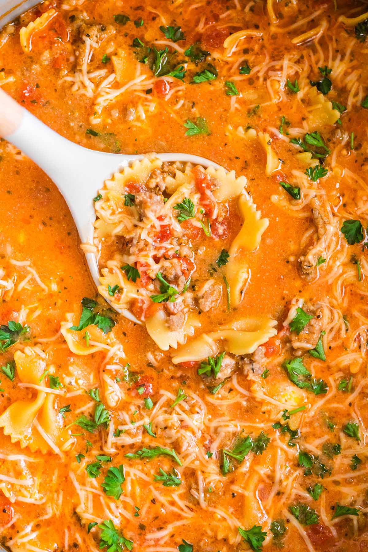 Close-up of lasagna soup in a pot, with a ladle holding bowtie pasta, sausage, and parsley.