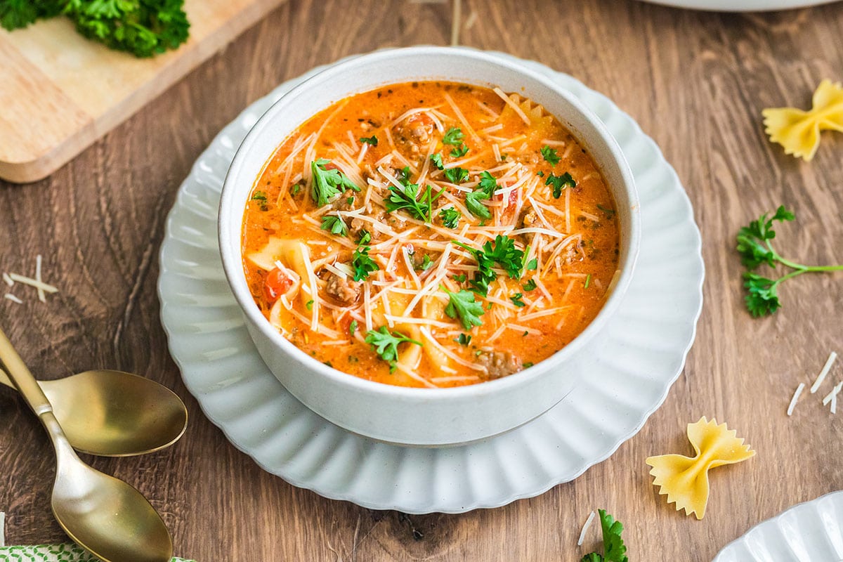 Lasagna soup topped with parsley and cheese in a white bowl on a wooden table.