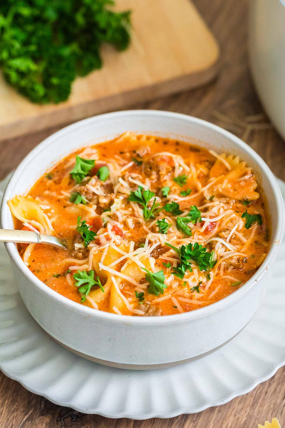 White bowl of lasagna soup garnished with parsley and cheese, served on a plate.