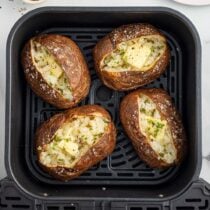 Air fryer baked potatoes split open with butter, salt, and chives inside the air fryer basket.