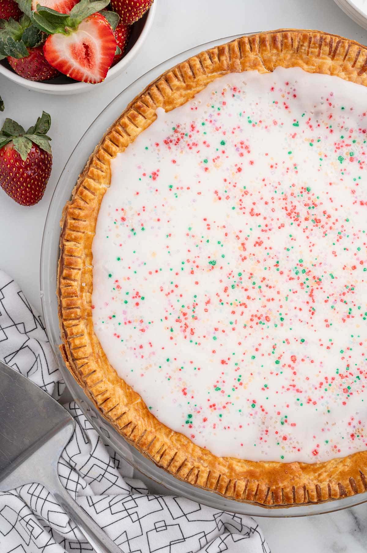 Strawberry Pop Tart Pie topped with icing and colorful sprinkles, with fresh strawberries on the side.