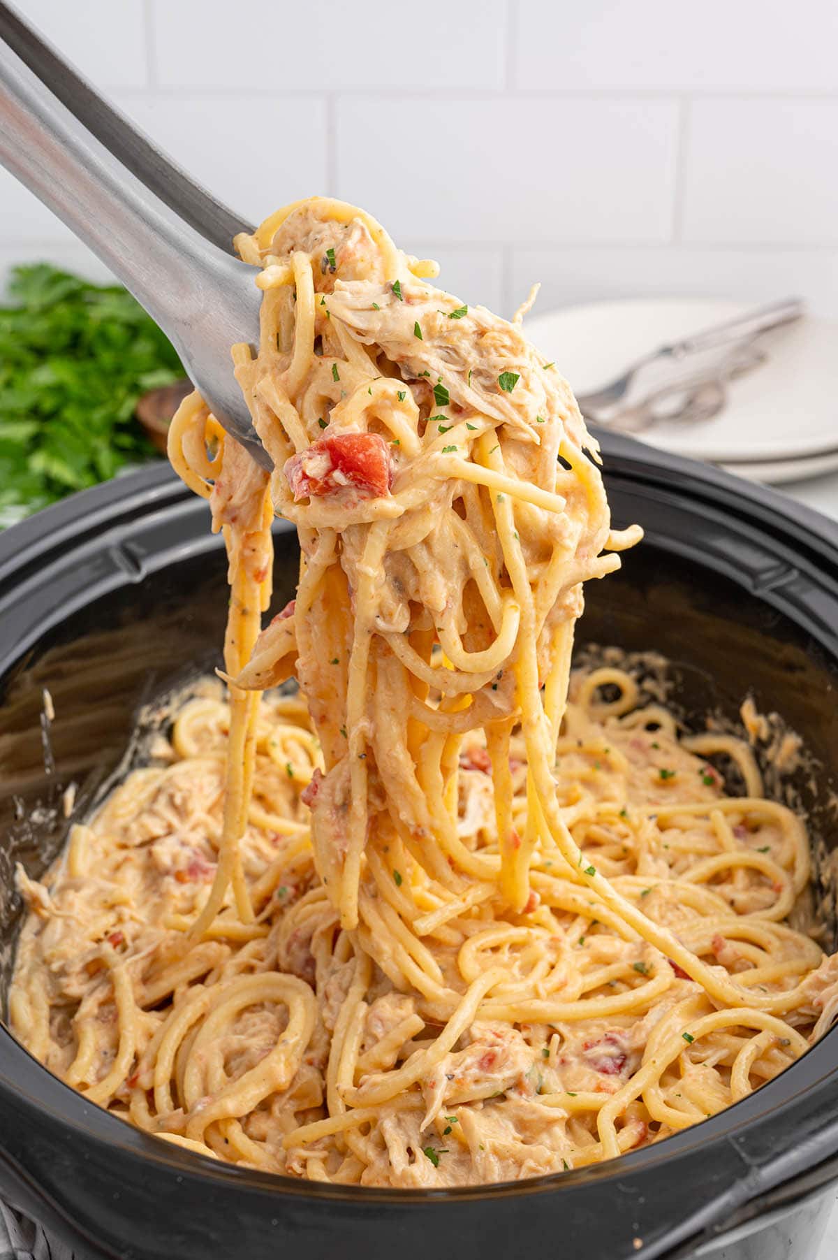 Tongs lifting creamy chicken spaghetti from a slow cooker.