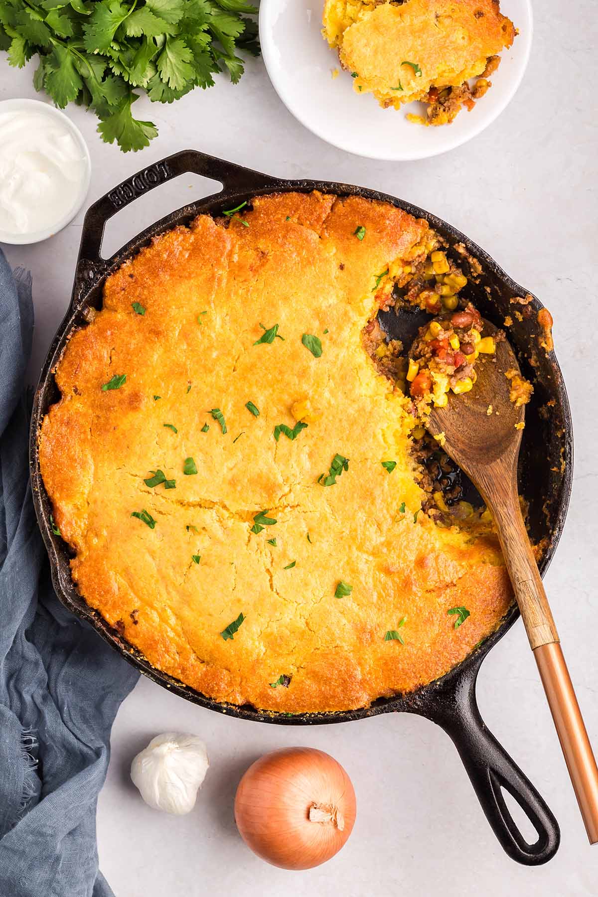 Cornbread cowboy casserole in a skillet with a wooden spoon and fresh toppings nearby.