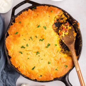 Cornbread cowboy casserole in a cast-iron skillet with a wooden spoon scooping a portion.