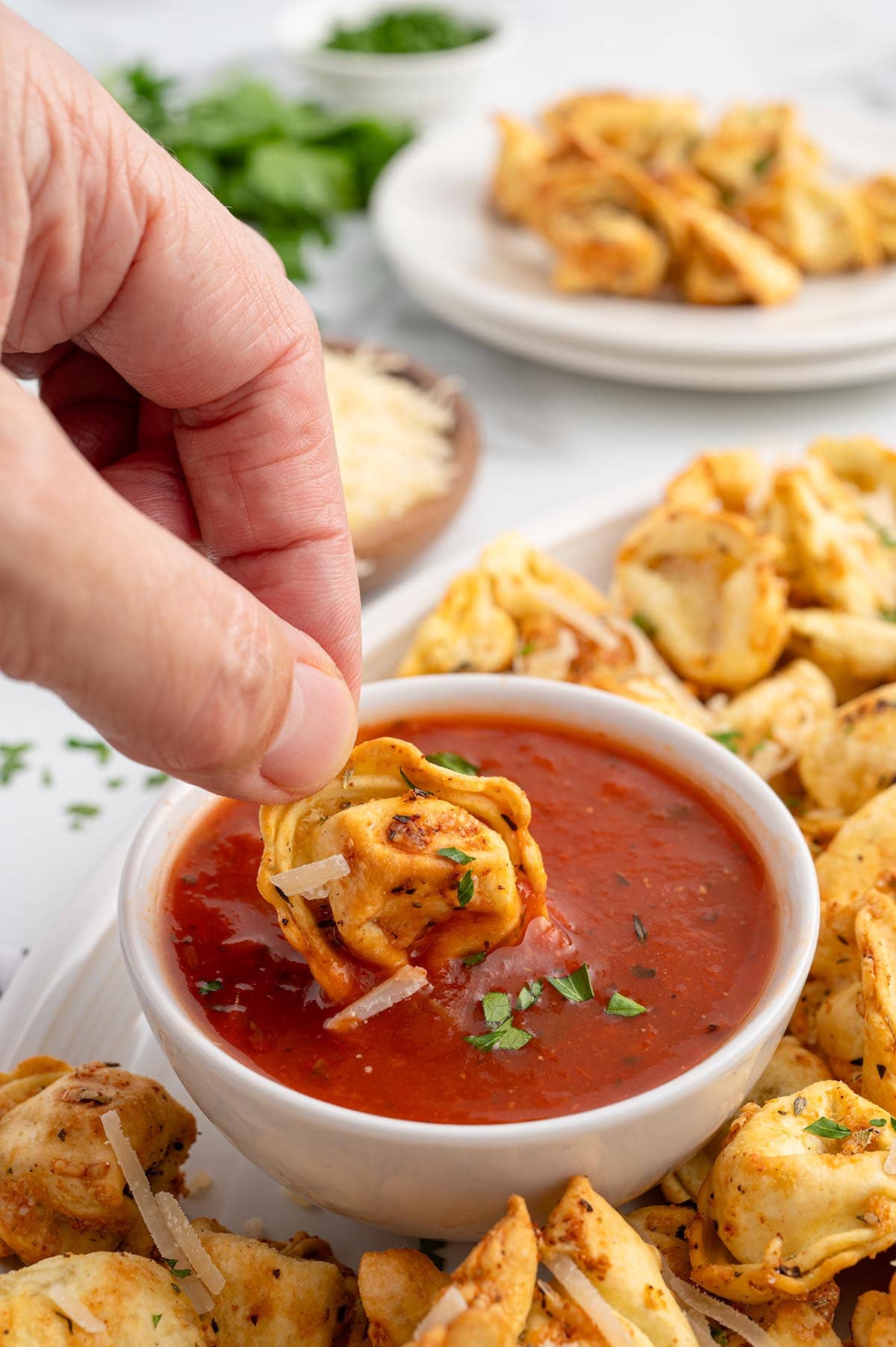 Hand dipping crispy air fryer tortellini into marinara sauce, topped with parmesan and parsley.