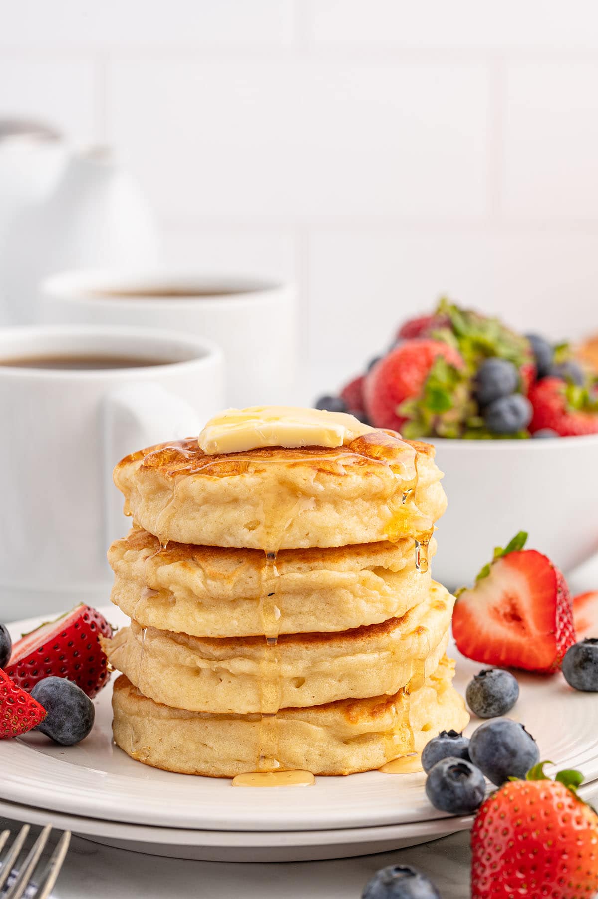 Stack of fluffy pancakes topped with butter and syrup, surrounded by fresh strawberries and blueberries.