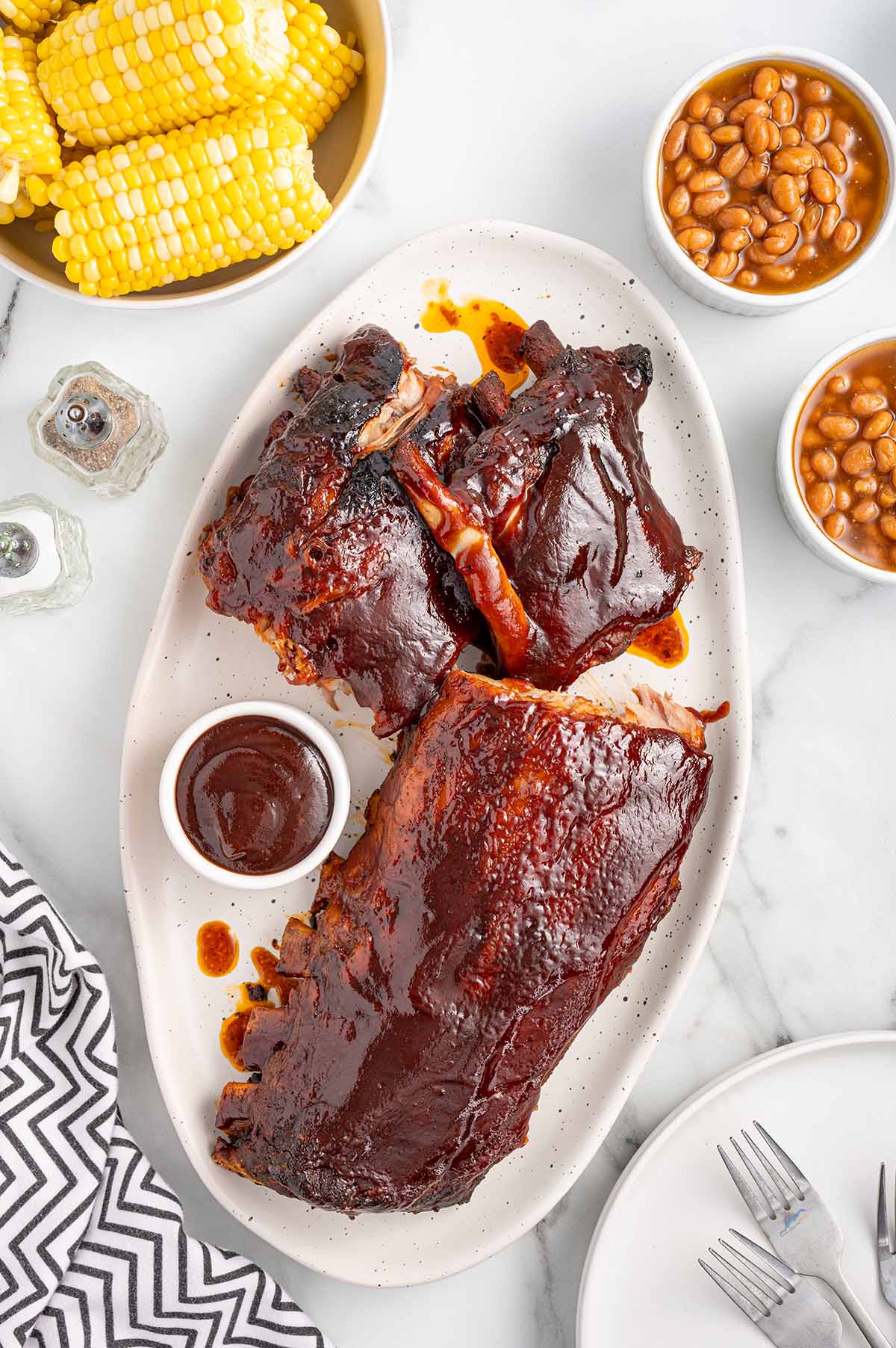 Slow Cooker Baby Back Ribs with barbecue sauce, corn, and baked beans on a white platter.