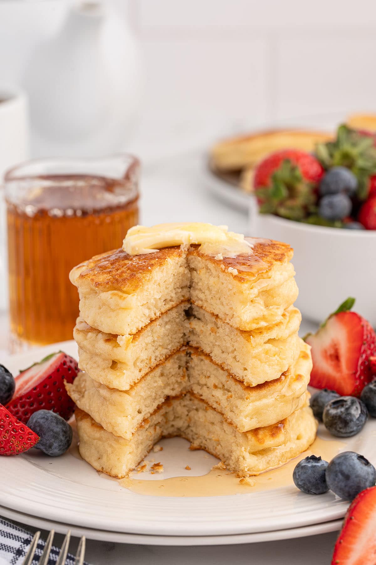 Stack of fluffy pancakes with a slice removed, topped with butter and syrup, served with fresh strawberries and blueberries.