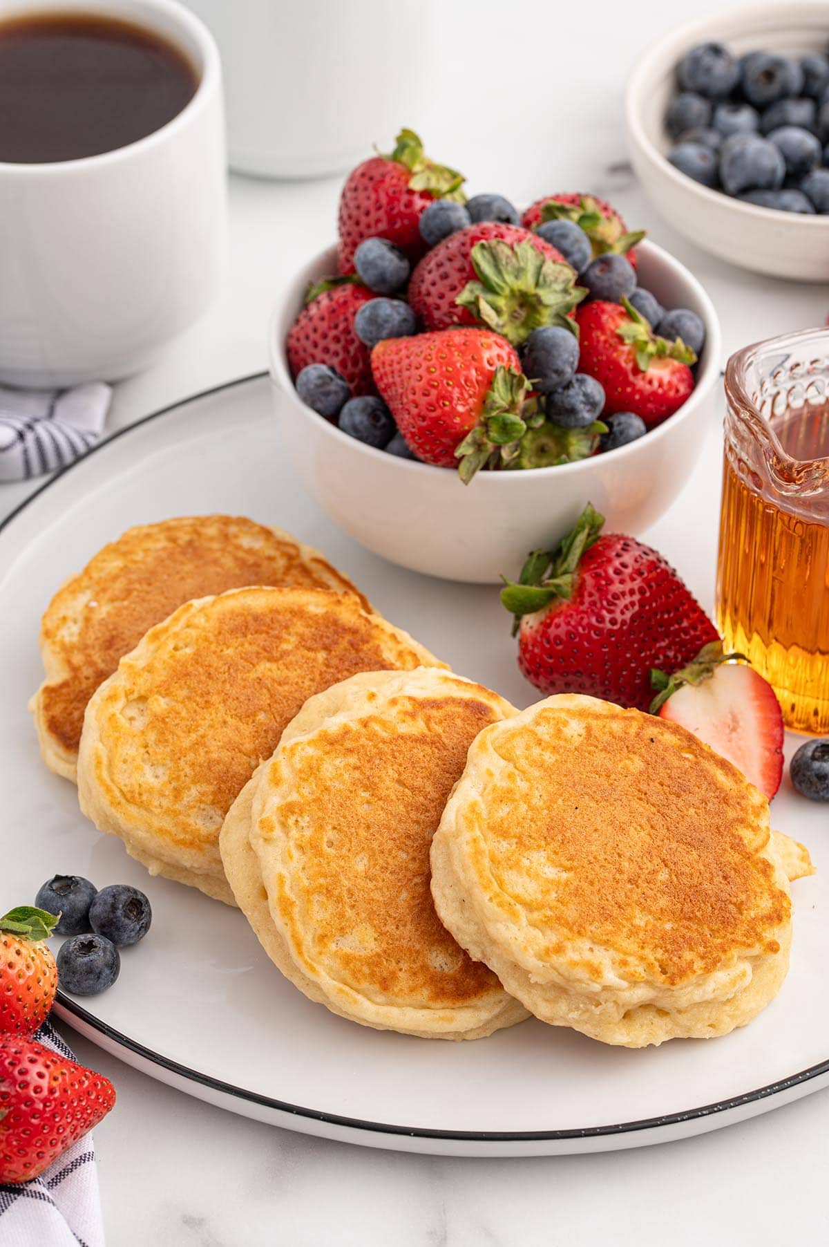 Plate of fluffy pancakes with a bowl of fresh strawberries and blueberries, syrup, and coffee in the background.