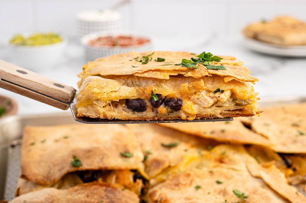 Spatula holding a slice of sheet pan chicken quesadilla with melted cheese, black beans, and crispy tortillas.