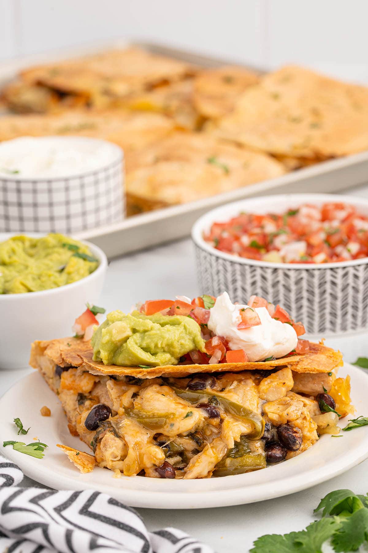 Slice of sheet pan chicken quesadilla on a plate, topped with guacamole, sour cream, and pico de gallo.