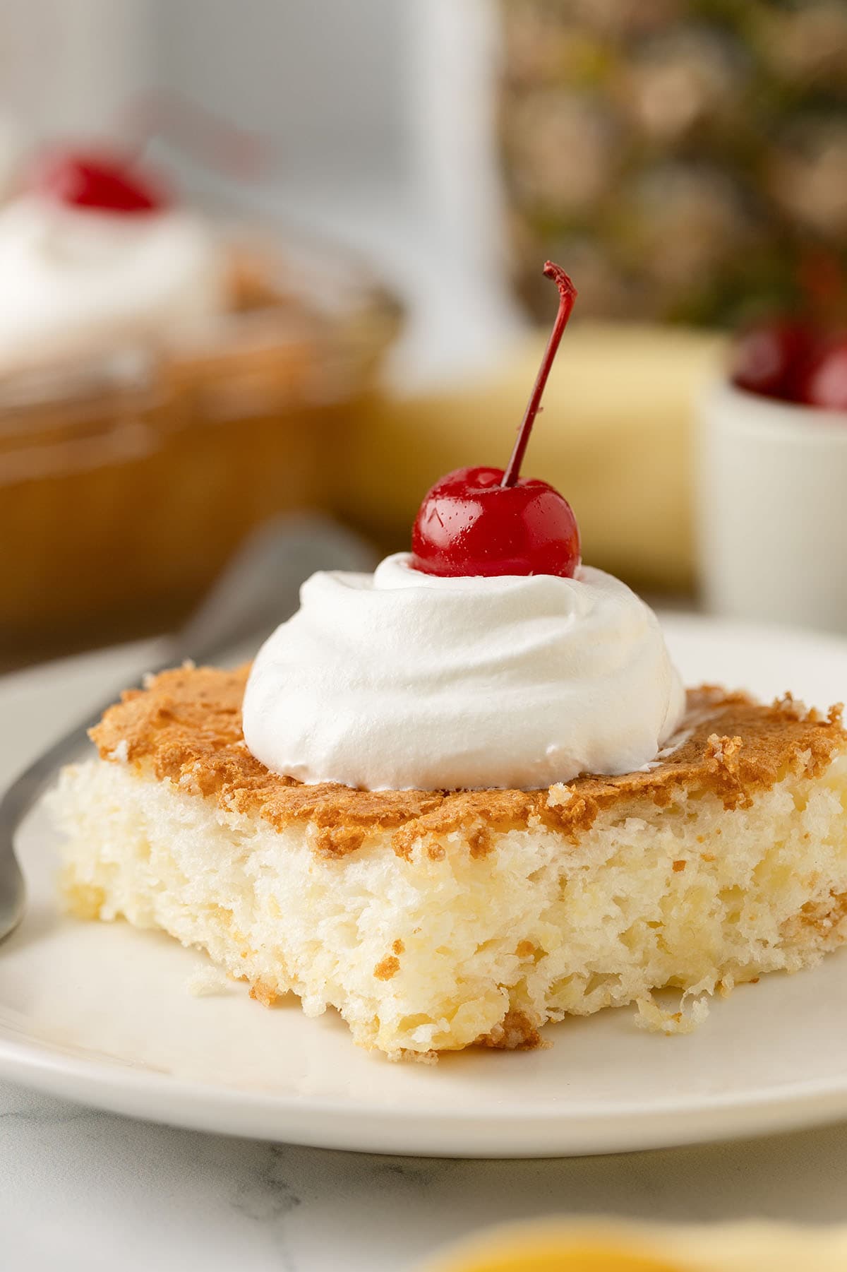 Close-up of a slice of pineapple angel food cake topped with whipped cream and a maraschino cherry.