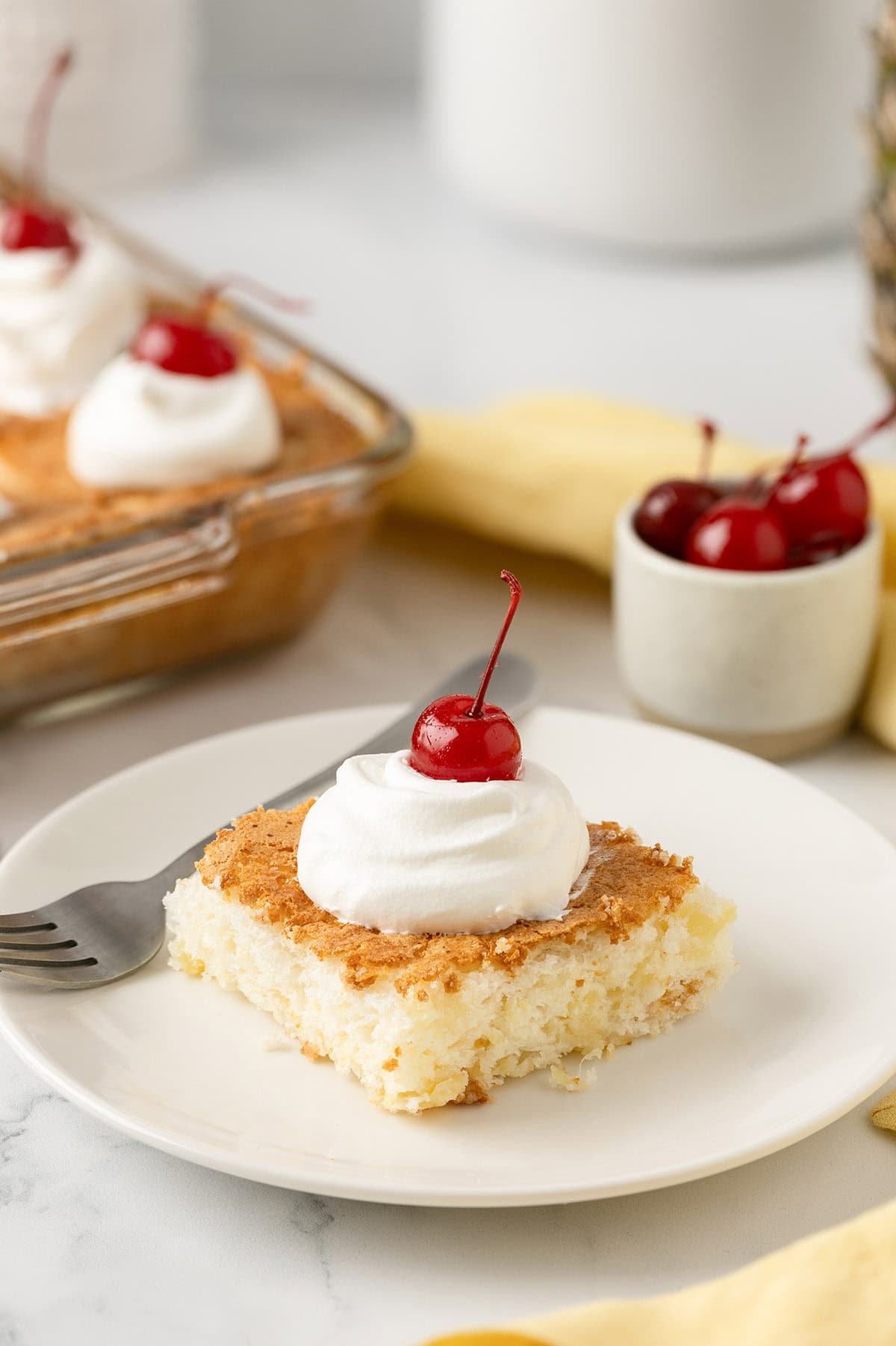Square slice of pineapple angel food cake topped with whipped cream and a cherry, served on a white plate.