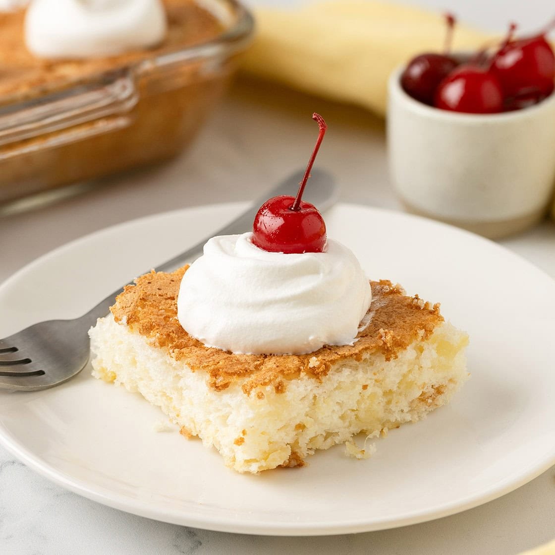 Slice of pineapple angel food cake topped with whipped cream and a cherry on a white plate.