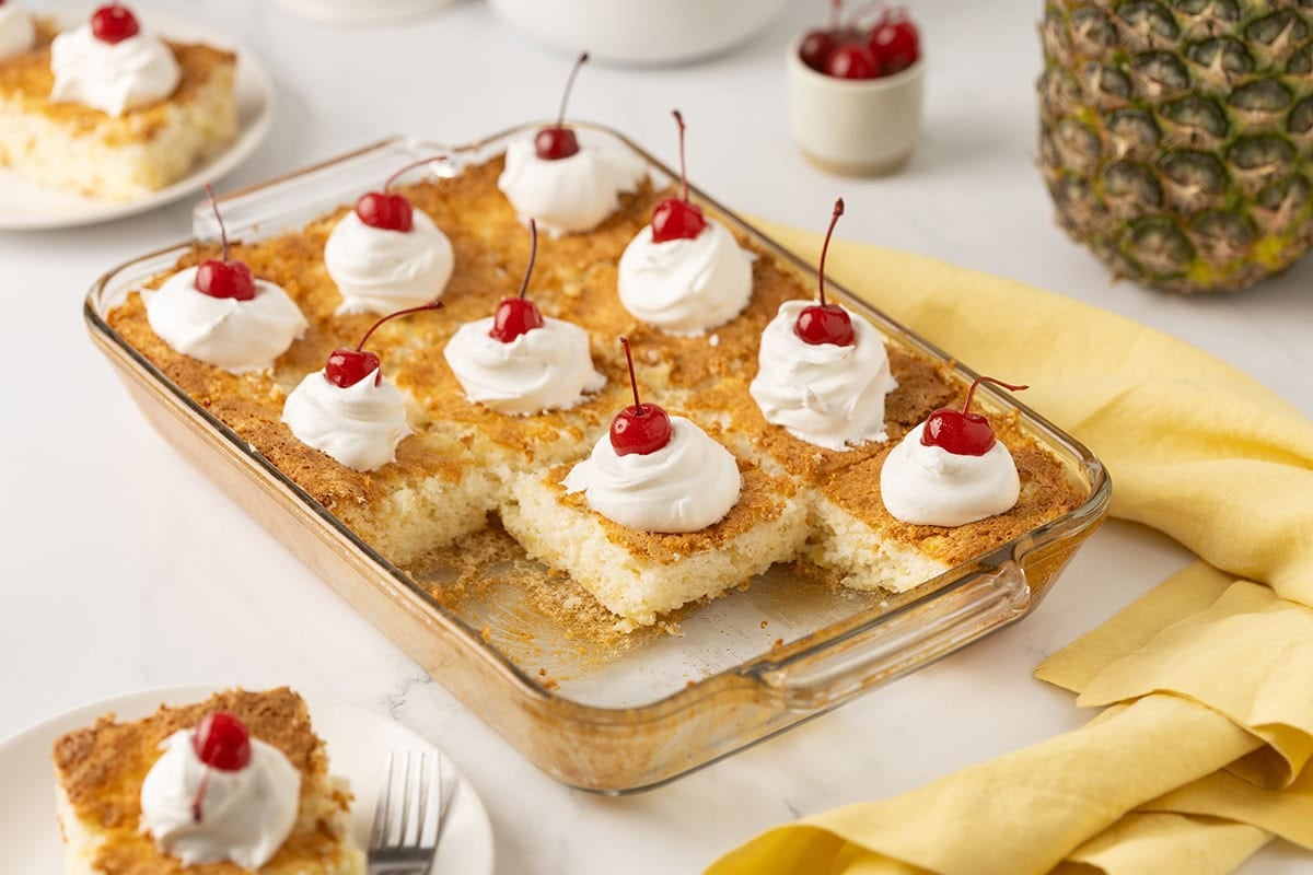 Glass baking dish filled with pineapple angel food cake squares, each topped with whipped cream and a maraschino cherry.