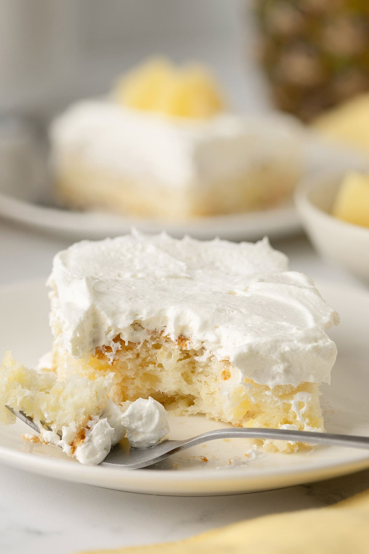 Slice of pineapple angel food cake topped with whipped cream, served on a white plate with a forkful taken out.