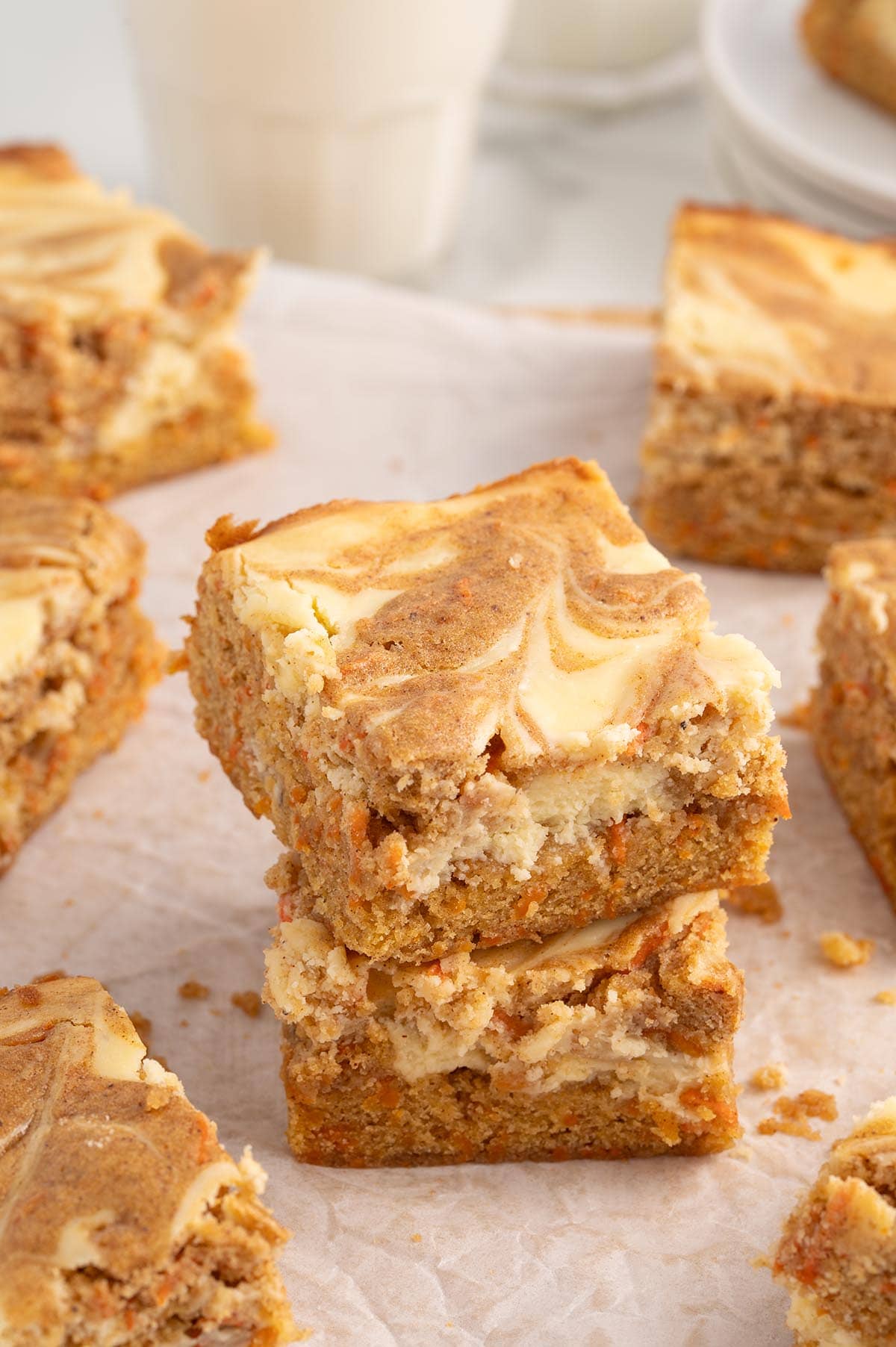 A stack of moist carrot cake bars with a rich cheesecake swirl, sitting on parchment paper with more bars in the background.