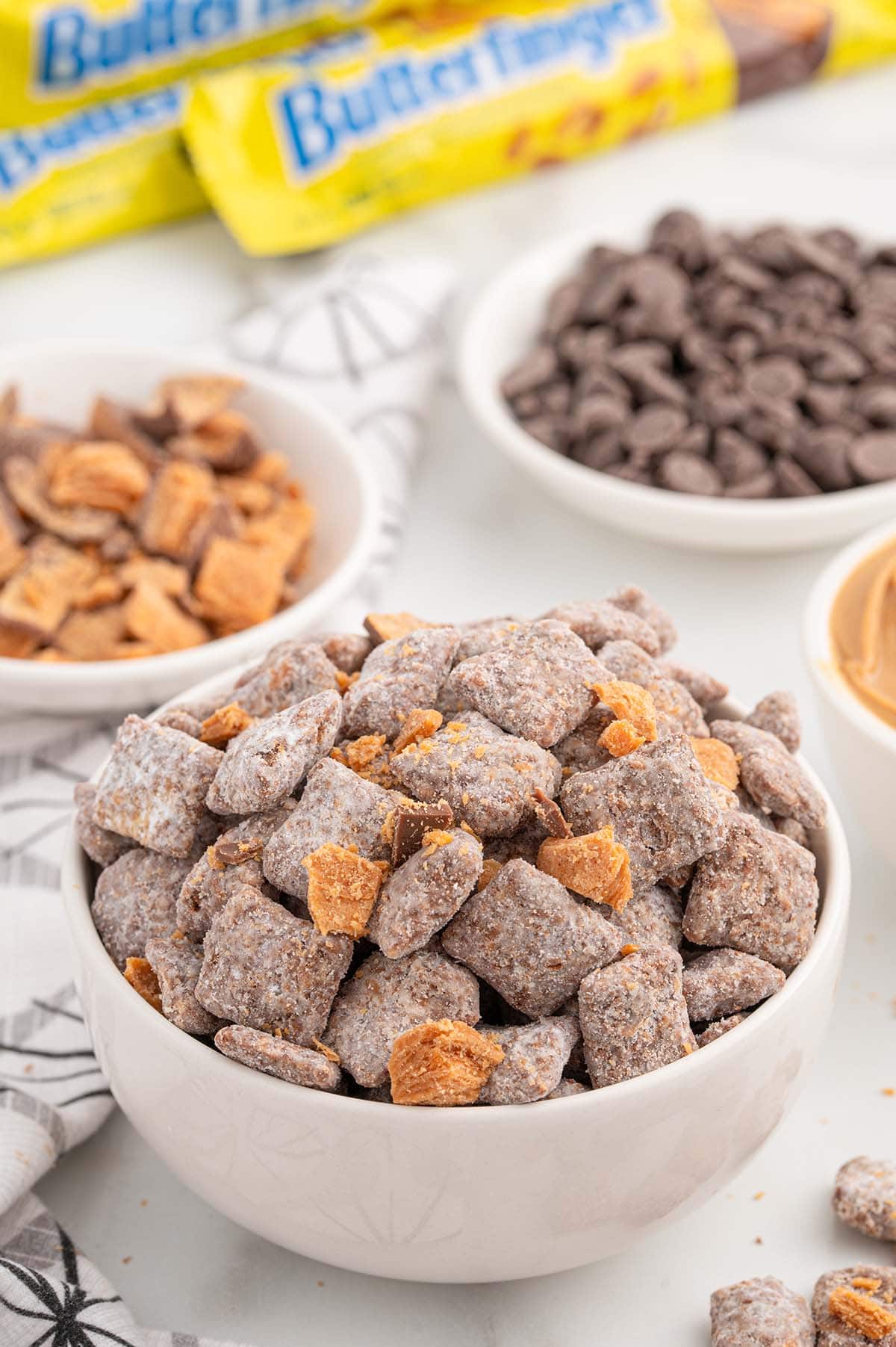 Butterfinger Puppy Chow in a bowl with chocolate chips and Butterfinger bars in the background.