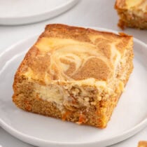 A square slice of moist carrot cake bar with a creamy cheesecake swirl on top, served on a white plate.