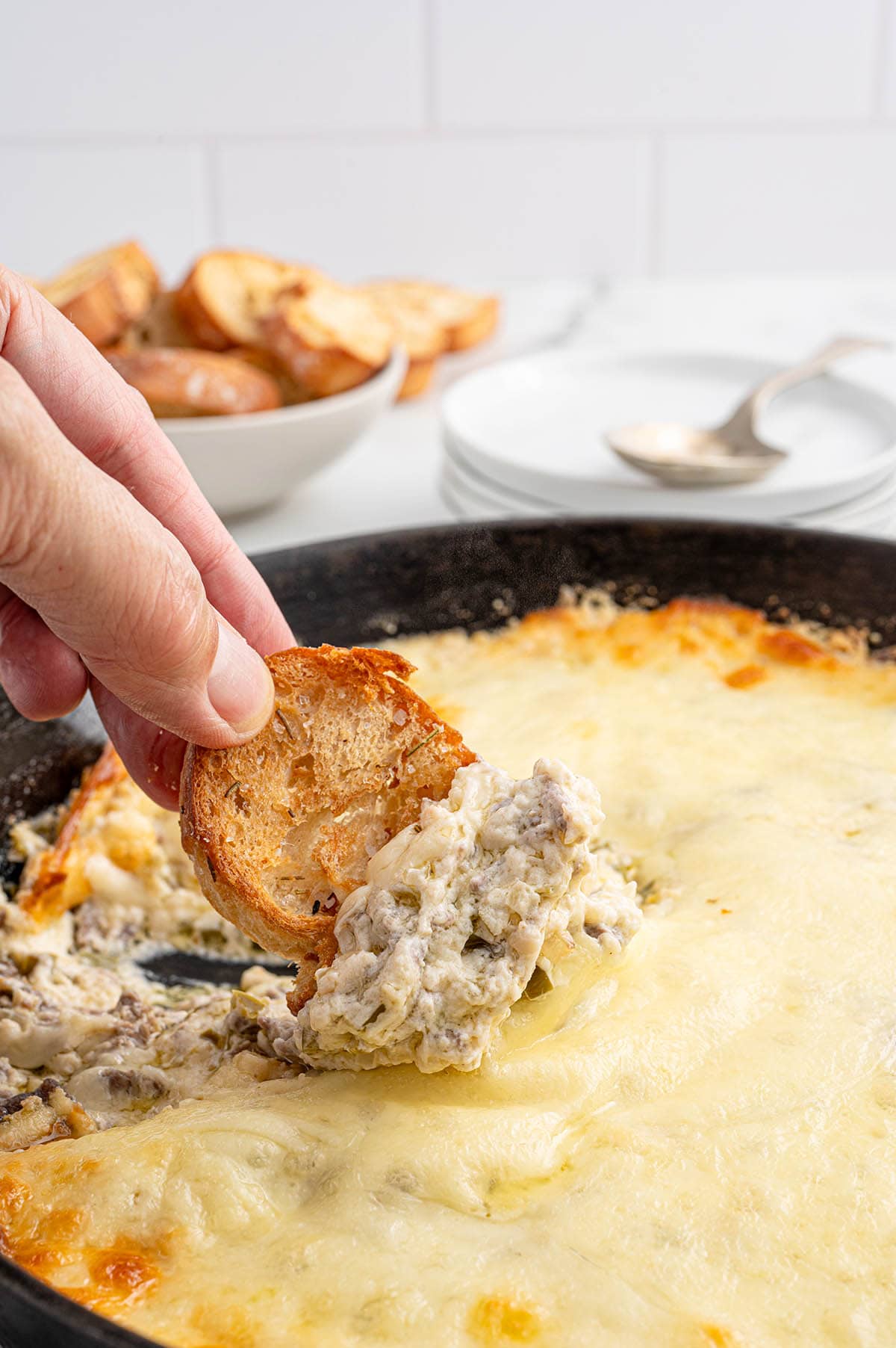 Toasted baguette slice dipped into Philly cheesesteak dip with gooey melted cheese in a skillet.