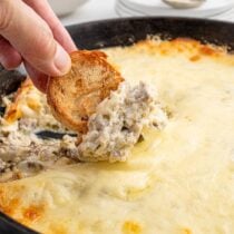 Philly cheesesteak dip scooped with a toasted baguette slice from a skillet with melted cheese.