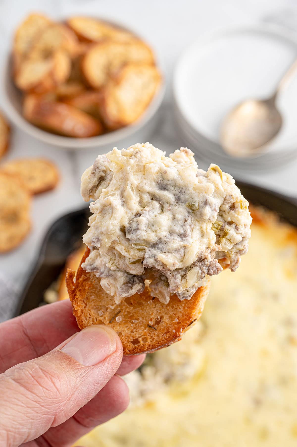 Philly cheesesteak dip scooped onto a toasted baguette slice, with a skillet of dip in the background.