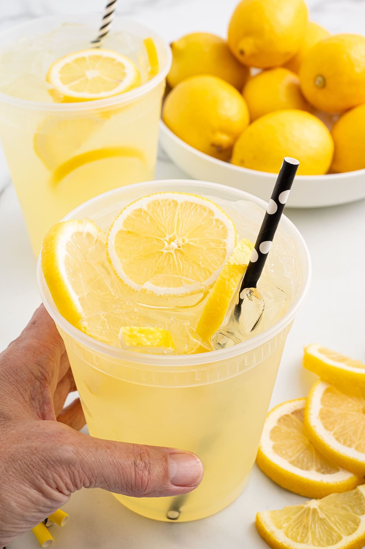 State Fair Lemonade with fresh lemon slices, ice, and a polka-dotted straw in a clear cup, held by hand.