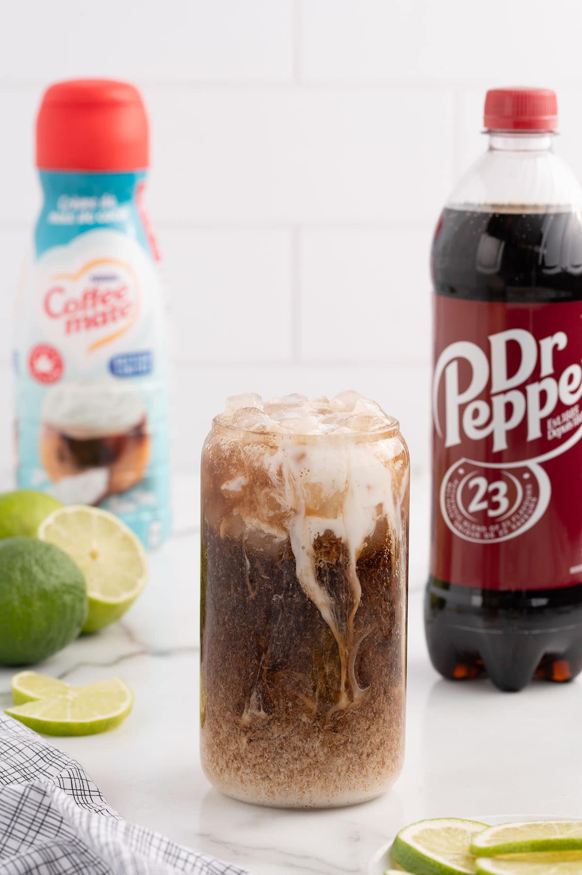 Glass of Dirty Dr Pepper with ice, lime slices, and a swirl of cream, surrounded by a bottle of Dr Pepper and coffee creamer.