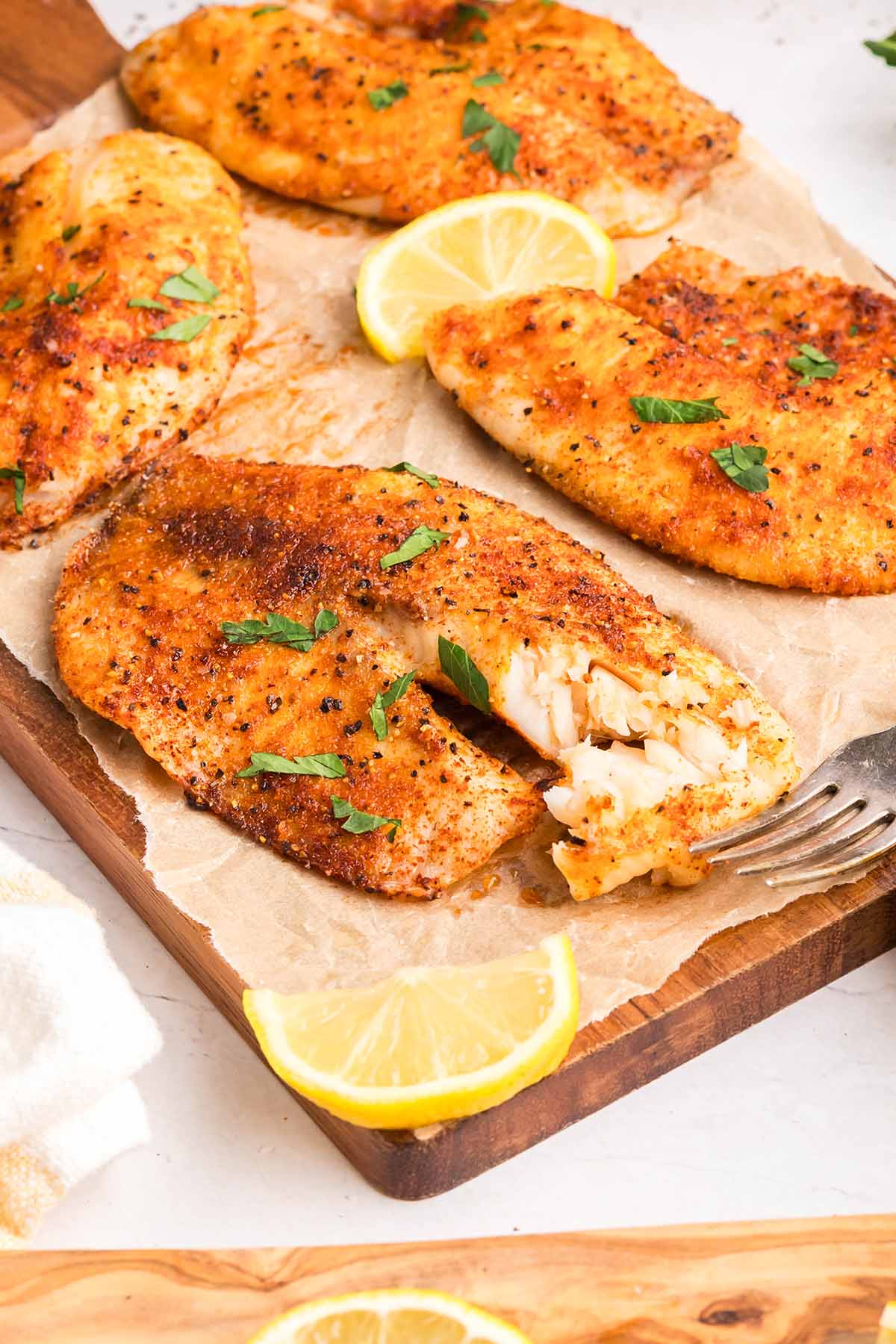 Fork flaking a piece of golden, seasoned air fryer tilapia fillet on parchment paper with lemon slices and parsley garnish.