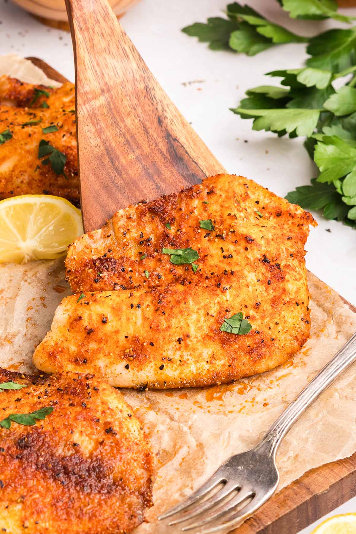 Golden, seasoned tilapia fillet being lifted with a wooden spatula, garnished with parsley and served with lemon.
