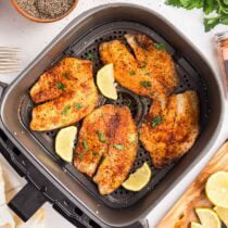 Raw tilapia fillets on parchment paper with seasoning ingredients including salt, pepper, paprika, onion powder, garlic powder, lemon, and parsley.