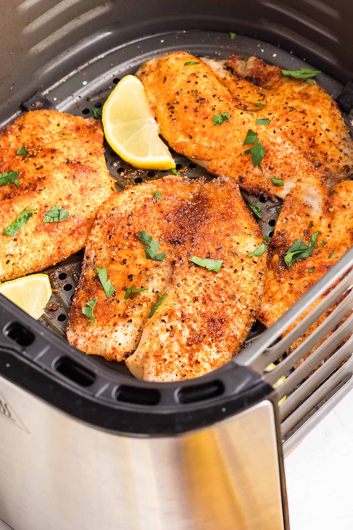 Seasoned tilapia fillets cooked in an air fryer basket, garnished with fresh parsley and lemon wedges.