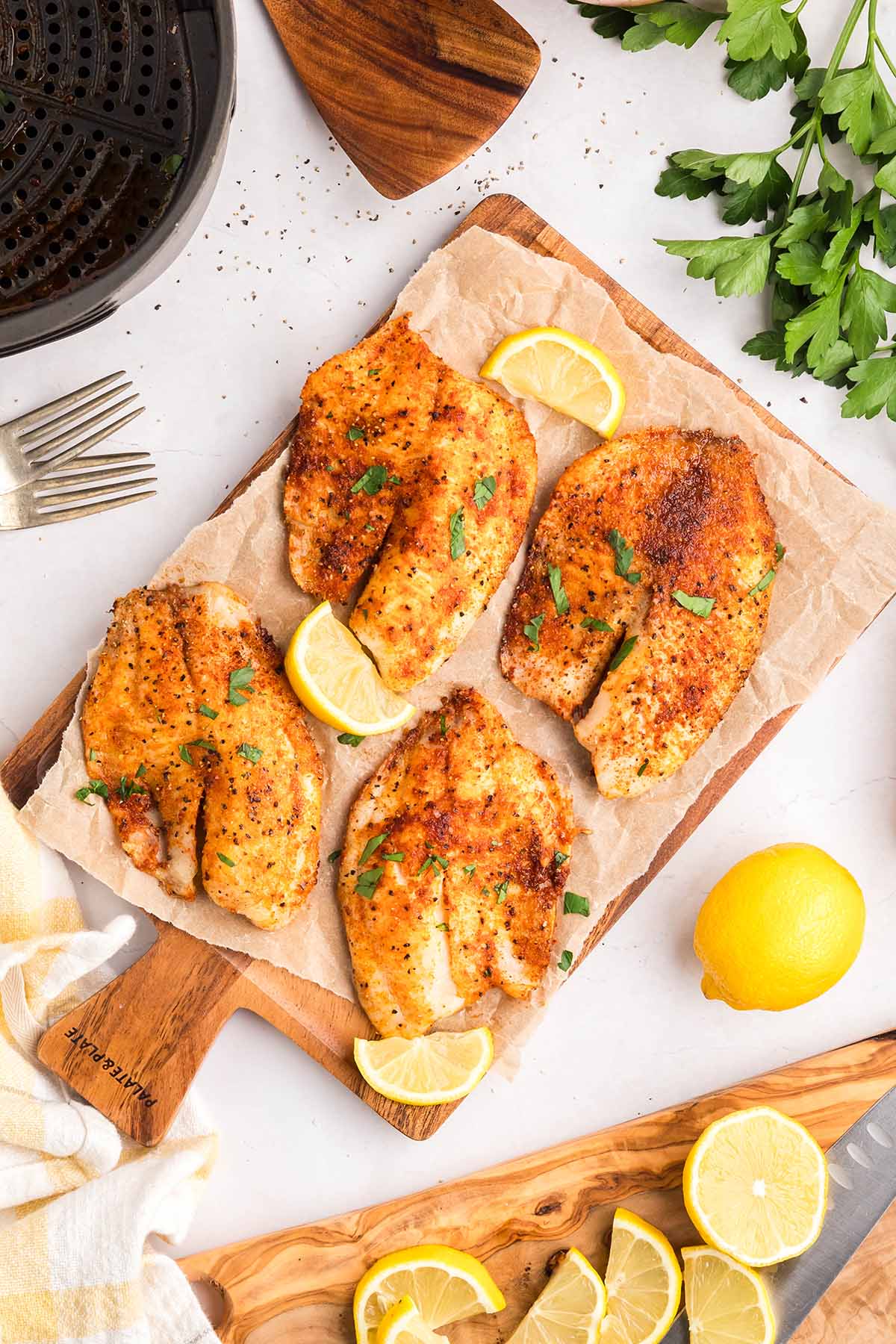 Four air-fried tilapia fillets on parchment-lined cutting board, garnished with parsley and lemon wedges, surrounded by fresh herbs and citrus.