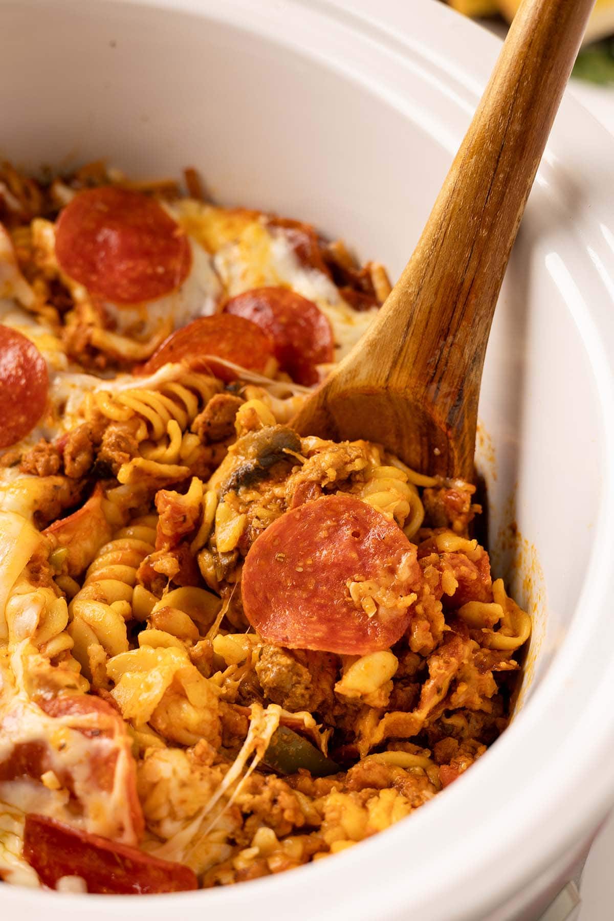Close-up of crock pot pizza pasta with rotini, melted cheese, sausage, and pepperoni, stirred with a wooden spoon.