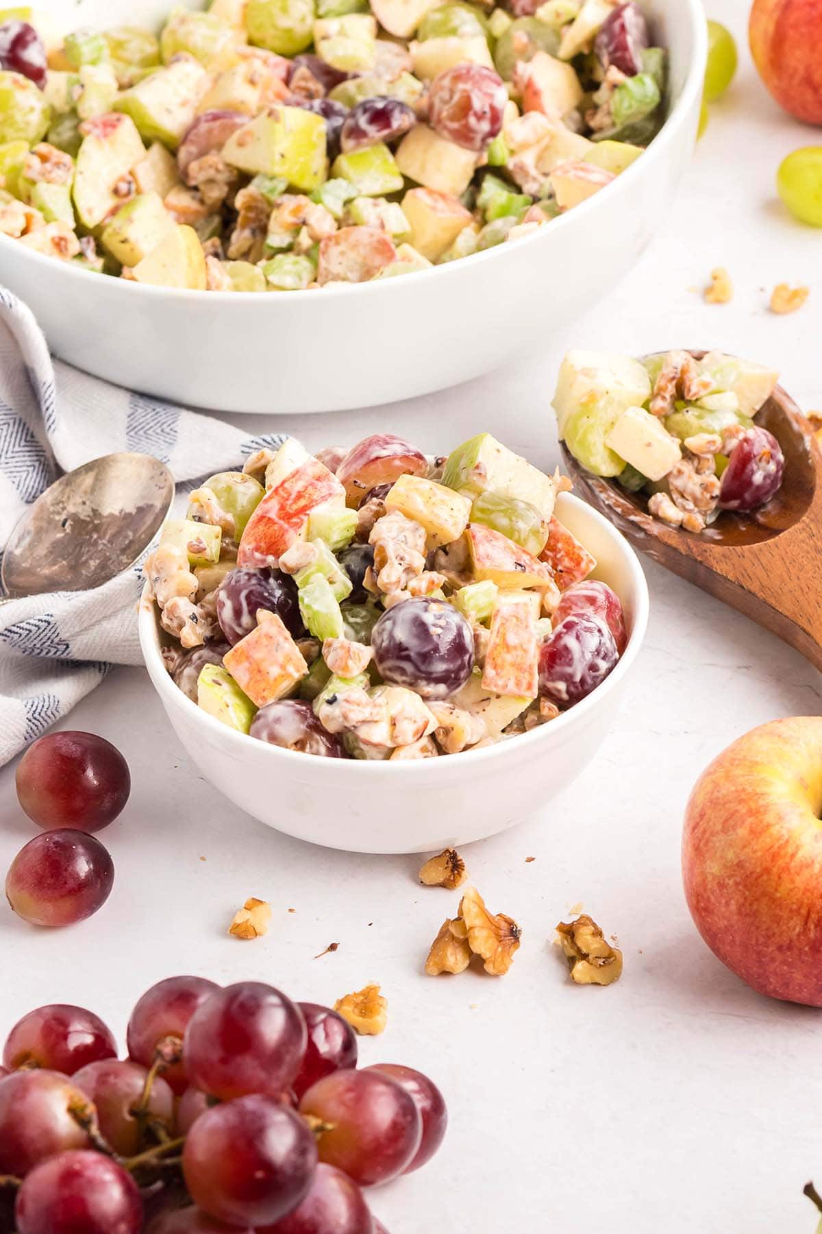 Small bowl of colorful Waldorf salad made with grapes, apples, celery, and walnuts, with ingredients scattered around and a larger serving bowl in the background.