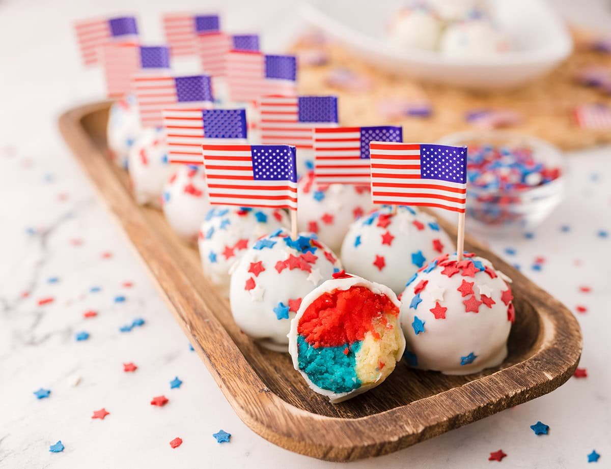 Patriotic cake balls decorated with red, white, and blue star sprinkles and mini American flags, displayed on a wooden tray.