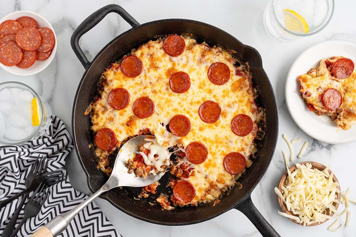 Crustless pepperoni pizza in a cast iron skillet with a slice served on a plate and shredded cheese on the side.