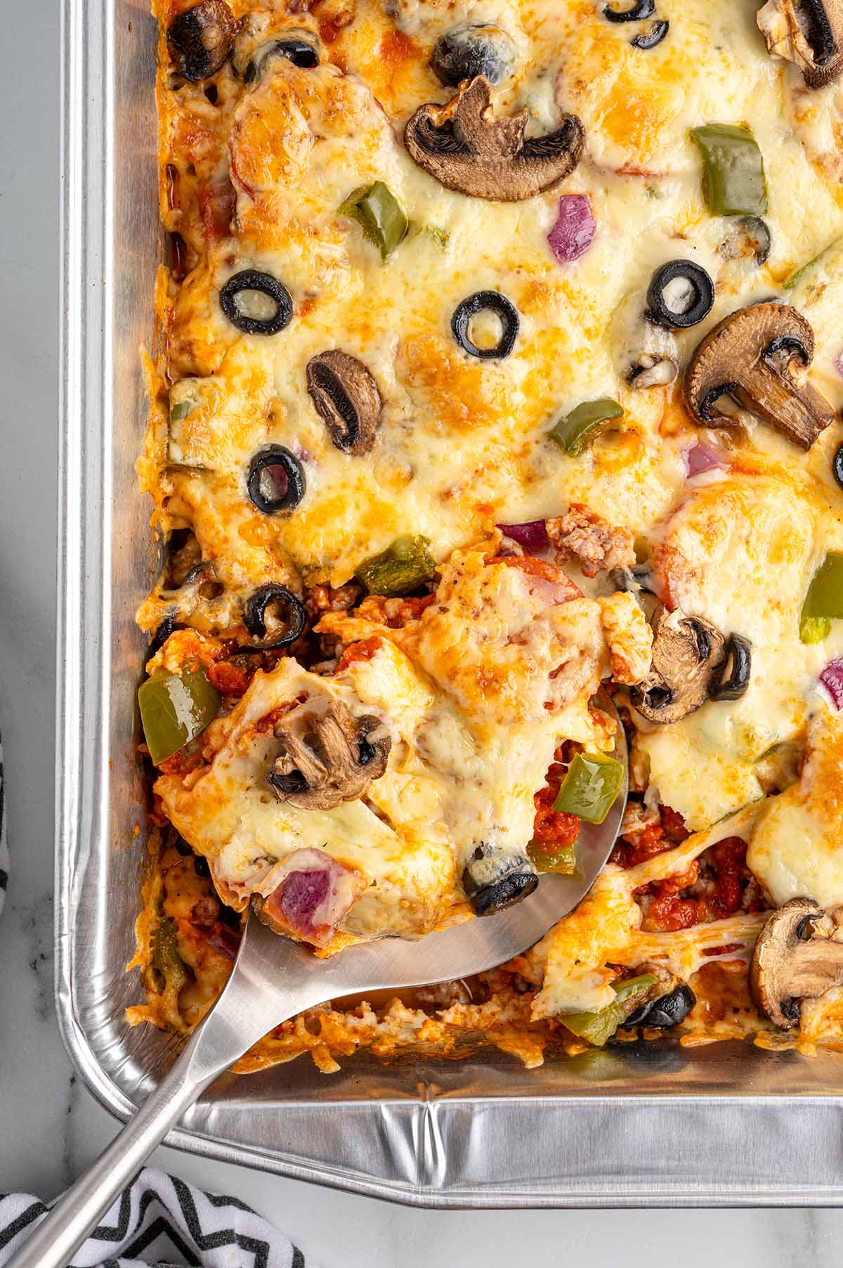 Slice of crustless supreme pizza with mushrooms, olives, green peppers, and melted cheese being served from a baking pan.