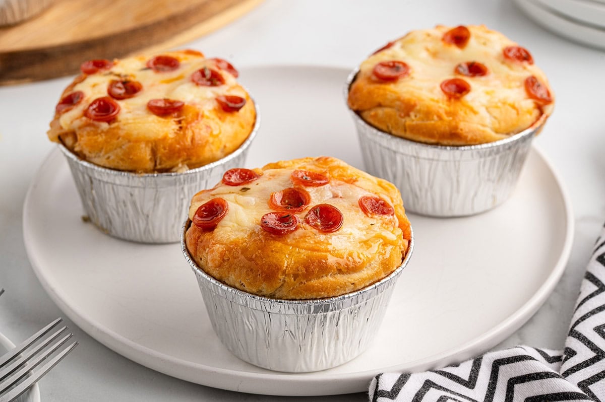 Three mini pizza pot pies in foil cups on a white plate, topped with melted mozzarella cheese and mini pepperoni slices.