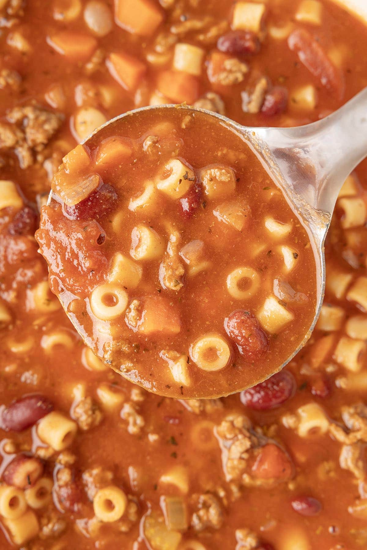 Close-up of a ladle filled with pasta e fagioli soup featuring ditalini pasta, beans, ground beef, carrots, and onions in a rich tomato broth.