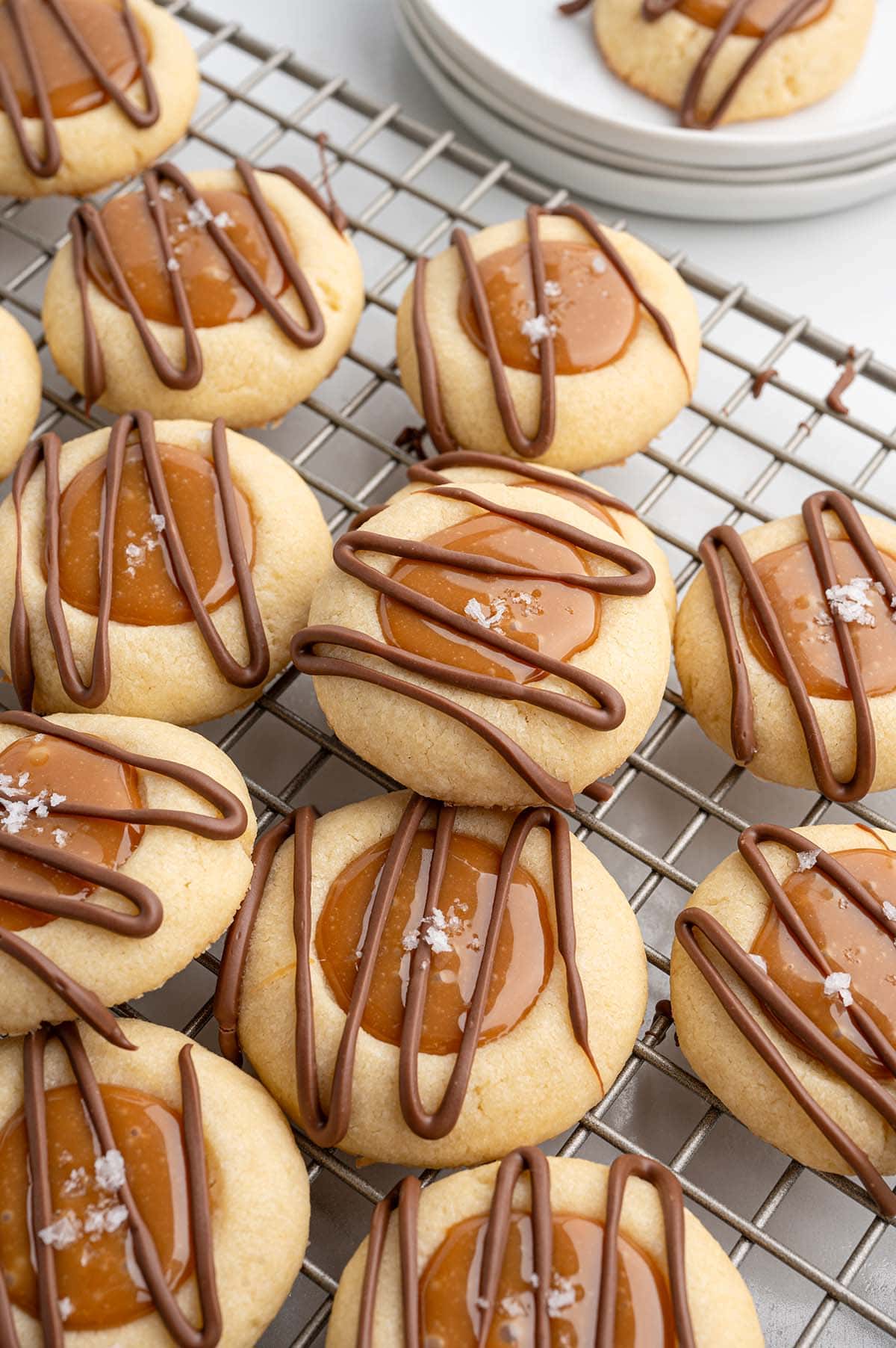 Close-up of golden Twix thumbprint cookies topped with caramel and sea salt on a wire rack.