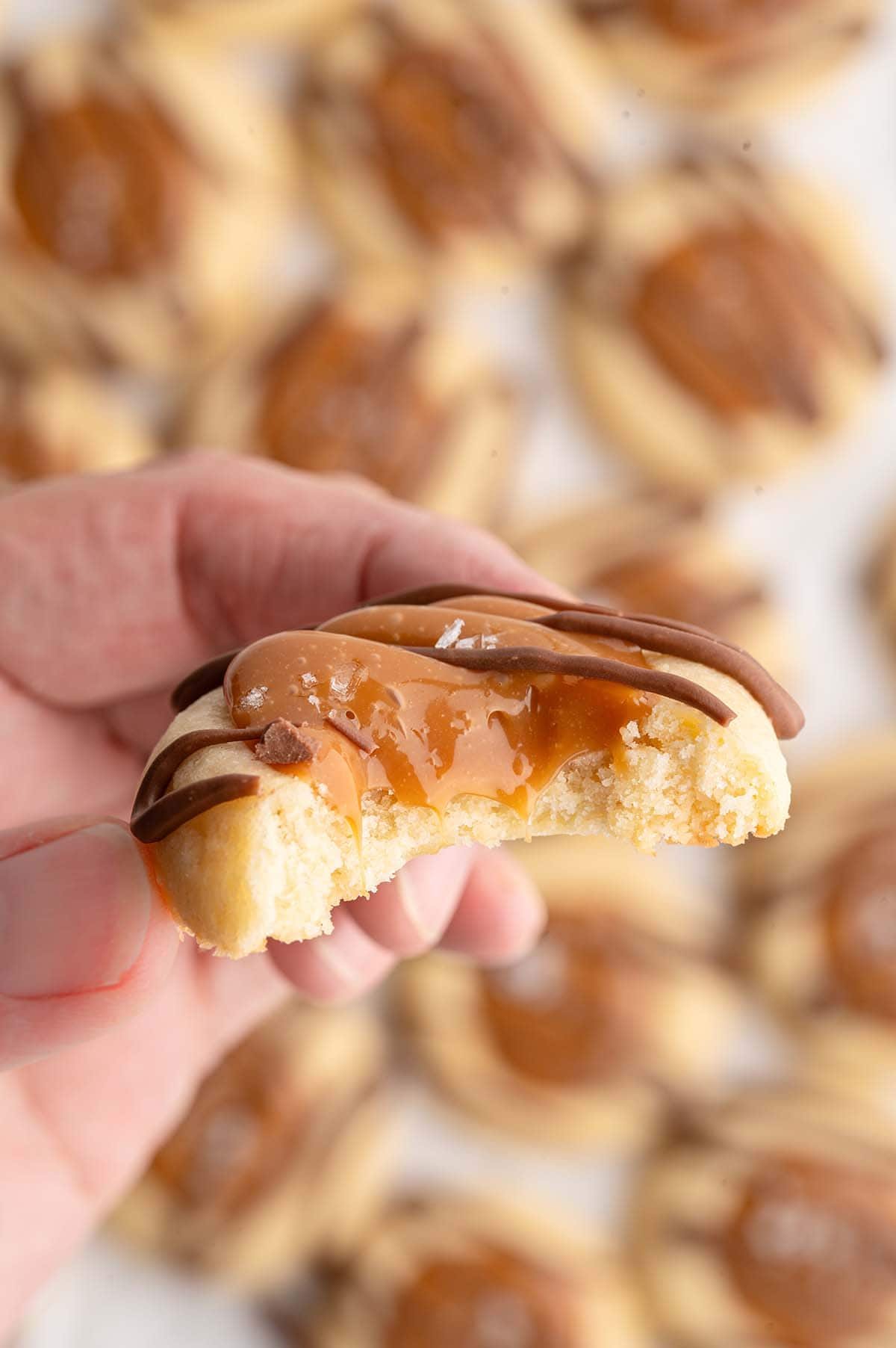 A hand holds a bitten Twix thumbprint cookies with gooey caramel and chocolate drizzle.