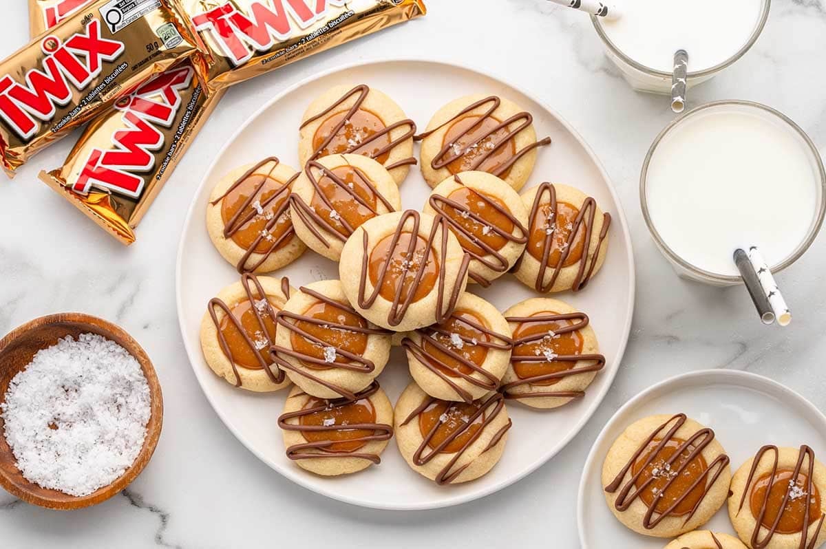 A plate of golden Twix thumbprint cookies topped with caramel and sea salt sits next to Twix bars and glasses of milk on a white table.