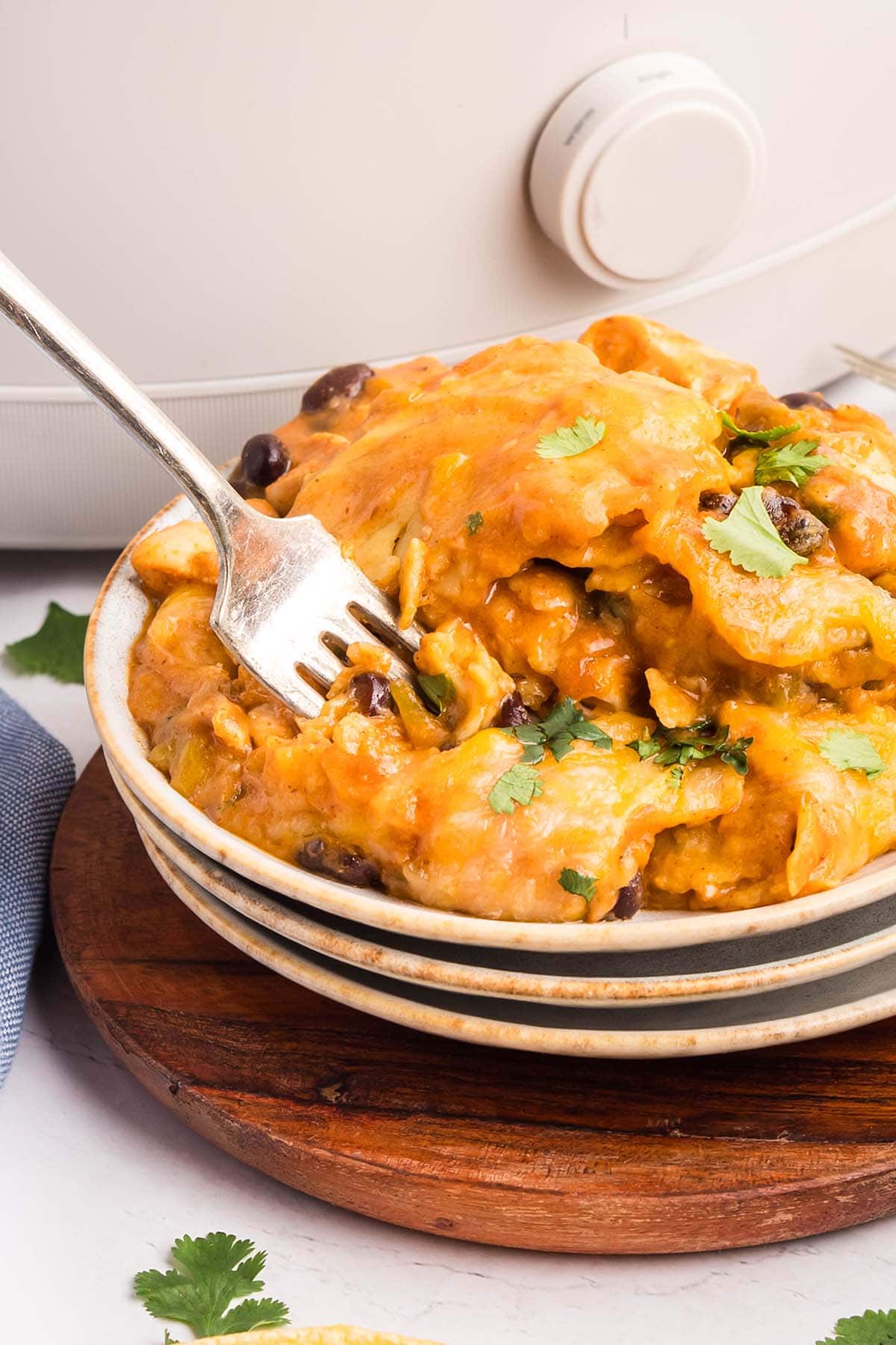 Close-up of a bowl of cheesy crockpot chicken enchilada casserole topped with cilantro, with a fork digging in and a slow cooker in the background.