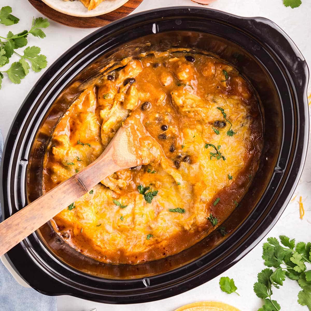 Cheesy crockpot chicken enchilada casserole topped with melted cheese and black beans, being stirred with a wooden spoon.
