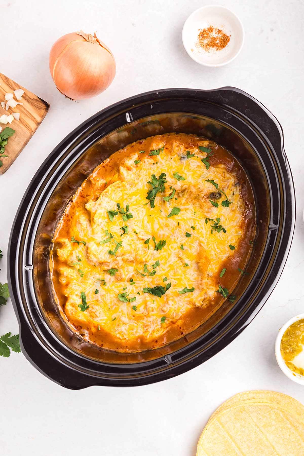 Overhead view of crockpot chicken enchilada casserole topped with melted cheese and fresh cilantro, surrounded by ingredients like onion, spices, and corn tortillas.